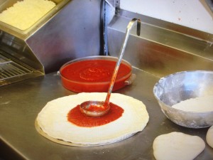 a ladle of pizza sauce on top of pizza dough near sauce and dough in bowls